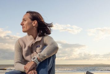 Kim Drenner, TCU alumna and head of environmental impact for Patagonia, sits barefoot on driftwood at a beach during golden hour, wearing a Patagonia sweater and jeans, with her hair blowing in the wind as she gazes thoughtfully toward the ocean. A wooden pier extends into the water in the background.