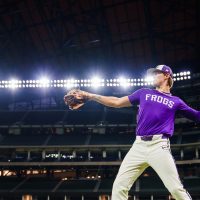 TCU baseball player in a purple top and light gray baseball pants warming up on the field at Globe Life Field under stadium lights.