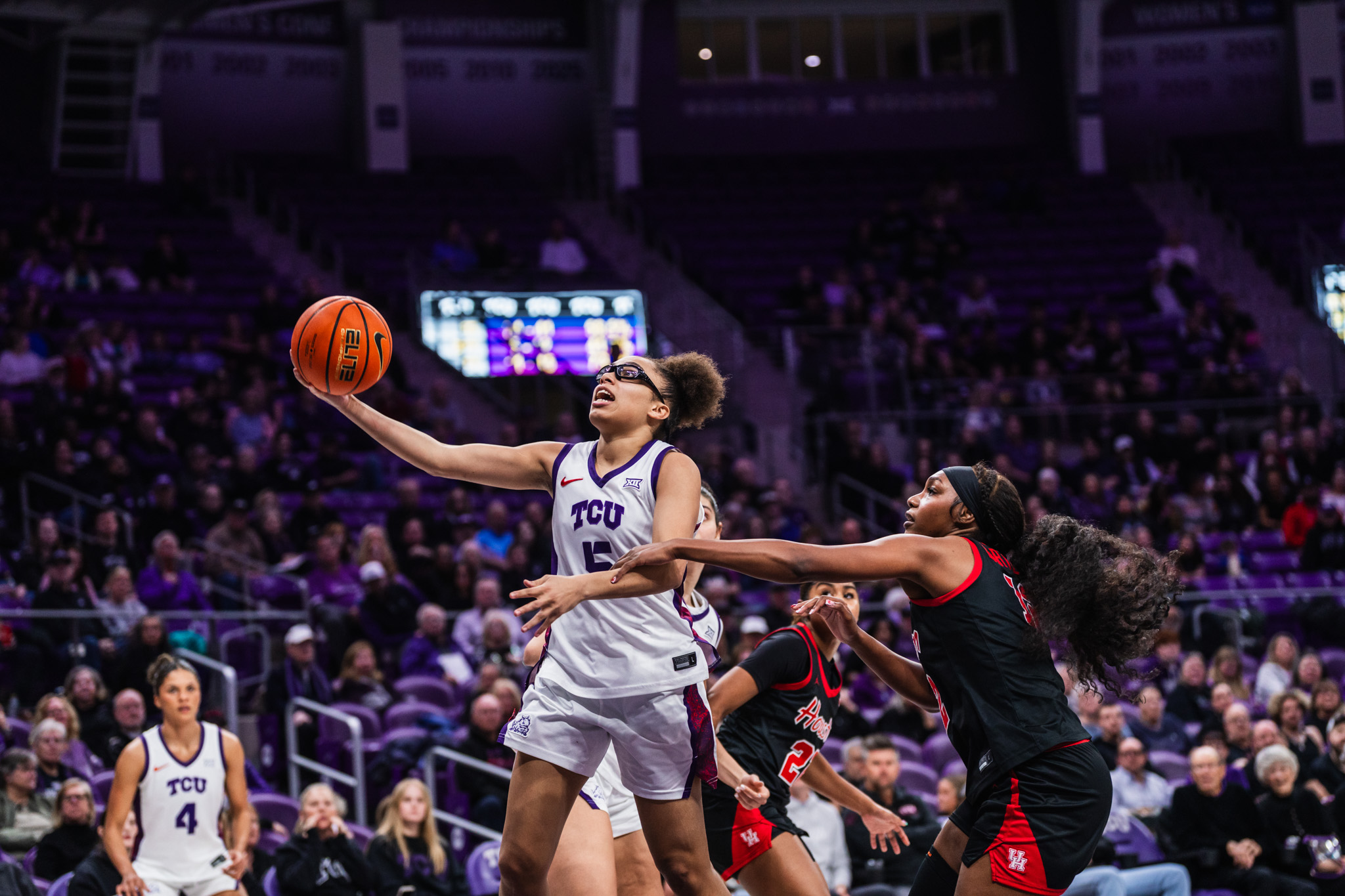 TCU guard Olivia Miles in white uniform extends for a layup while being defended by two Houston players in black and red uniforms at a packed home arena.