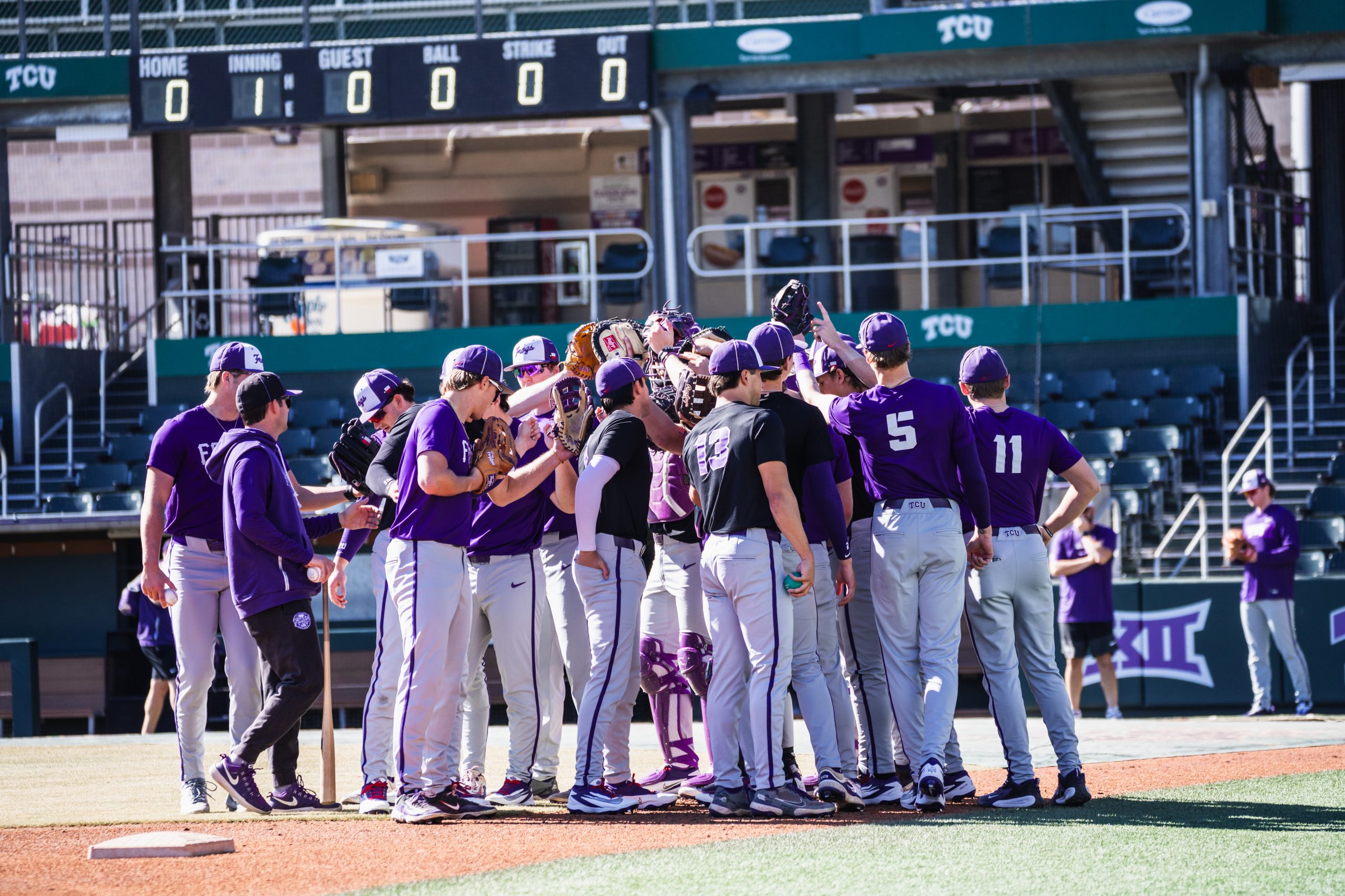 TCU baseball team huddles together on the field during practice at Lupton Stadium, with empty stands visible in the background.