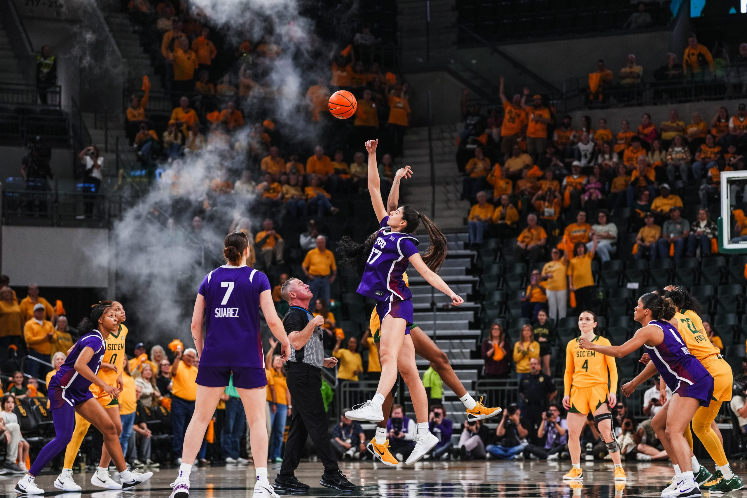 TCU's No. 17 goes up for a jump ball over a Baylor opponent during an NCAA basketball game at Baylor, with Marta Suárez (7) looking on and smoke billowing in the background as the gold-clad Baylor crowd fills the arena.