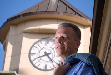 David Allen, the Luther A. Henderson University Chair in Leadership at the TCU Neeley School of Business, stands outdoors on a sunny day with a blazer slung over his left shoulder; a campus clock tower rises in the background.
