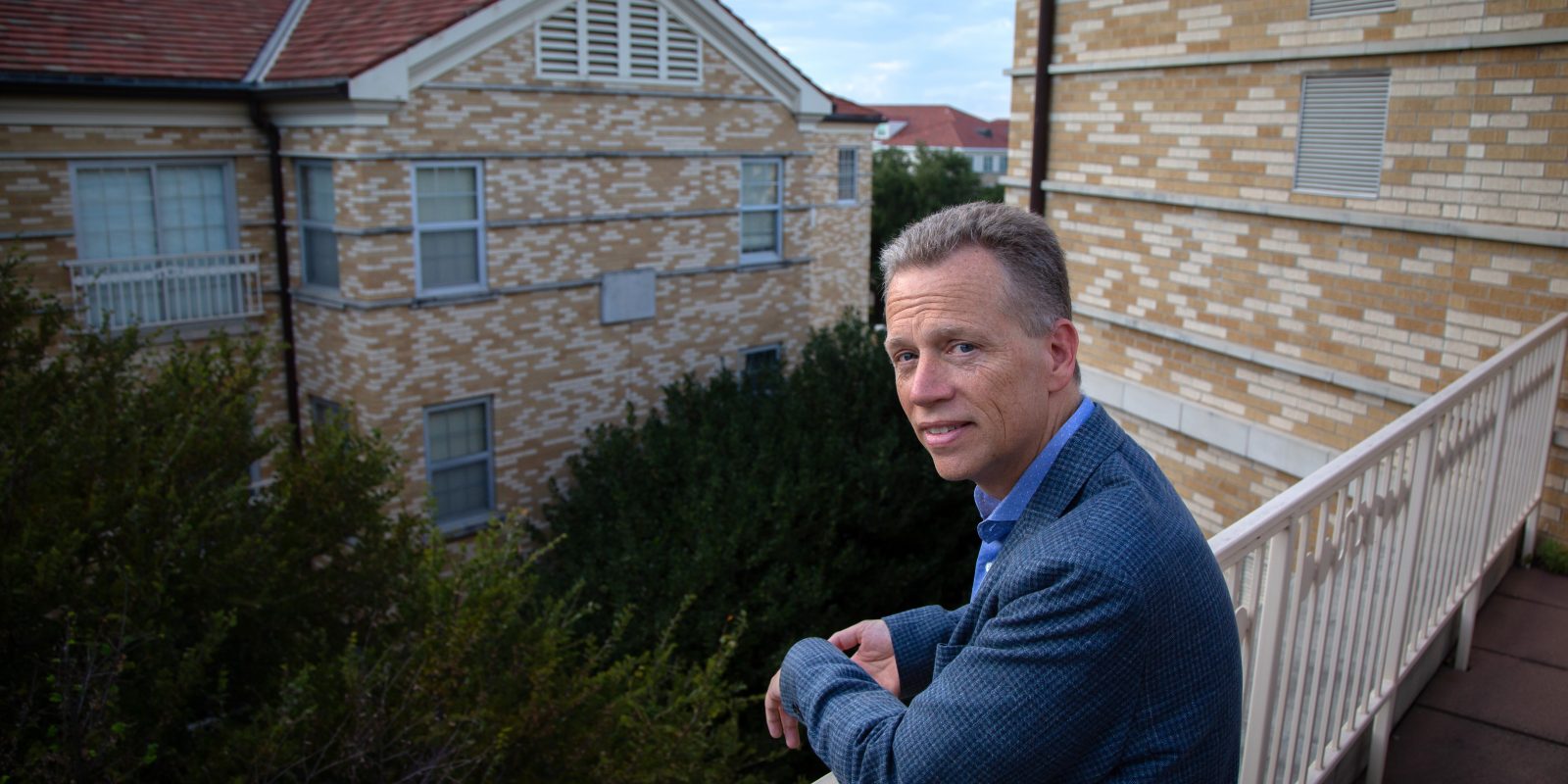 David Allen looks over his shoulder at the camera while wearing a dark blue sport coat and dress shirt, leaning against a railing with tan-brick buildings in the background.