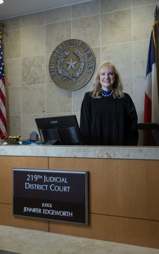 Judge Jennifer Edgeworth stands at the bench in her courtroom, wearing black judicial robes and a blue necklace. Behind her is the State of Texas seal mounted on a beige tiled wall, flanked by the American flag on the left and Texas flag on the right. A nameplate on the bench identifies the 219th Judicial District Court and Judge Jennifer Edgeworth.