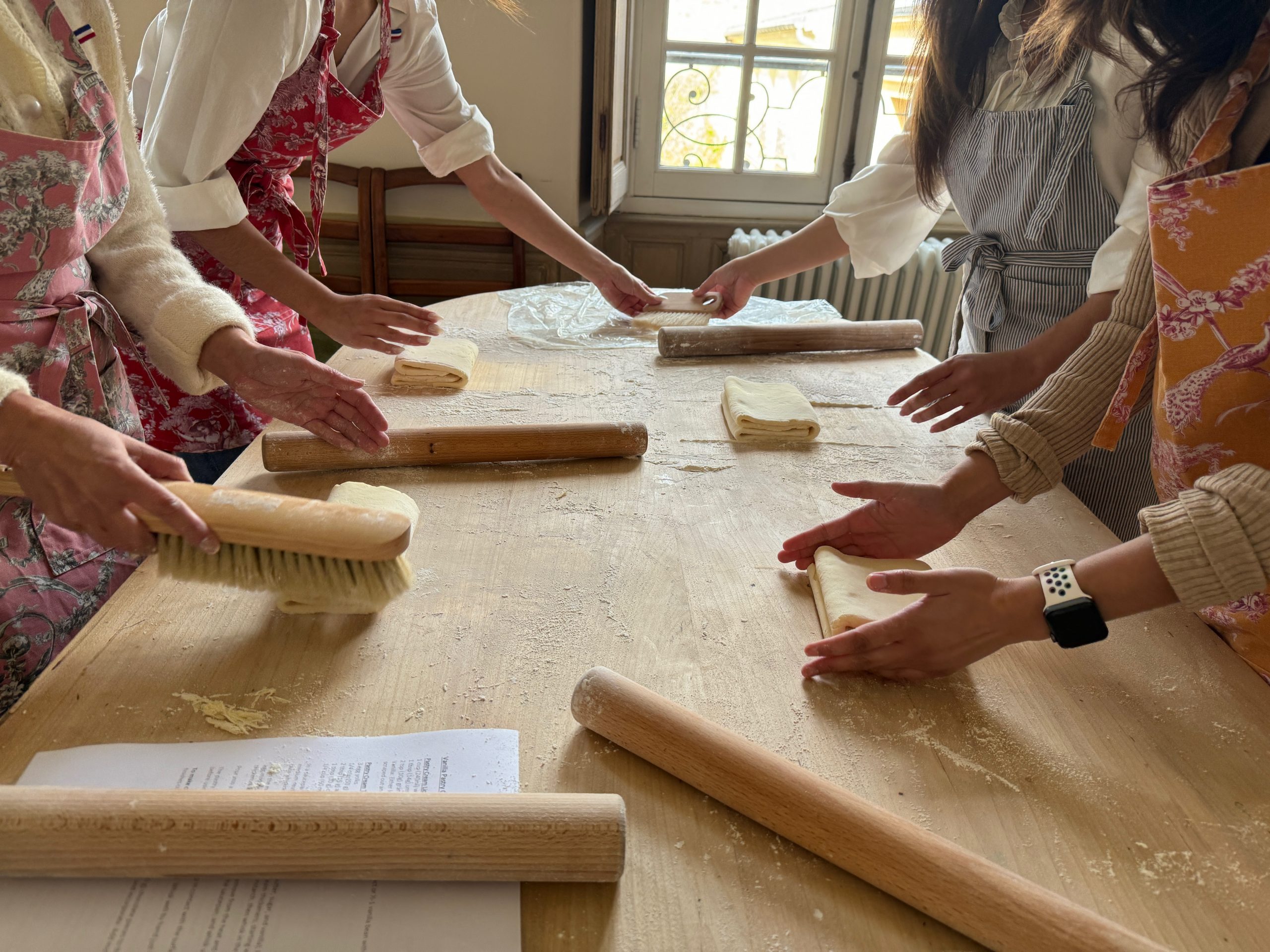 Several people in aprons work dough at a flour-dusted wooden table, with rolling pins and a pastry brush spread across the surface and a recipe sheet visible in the foreground.