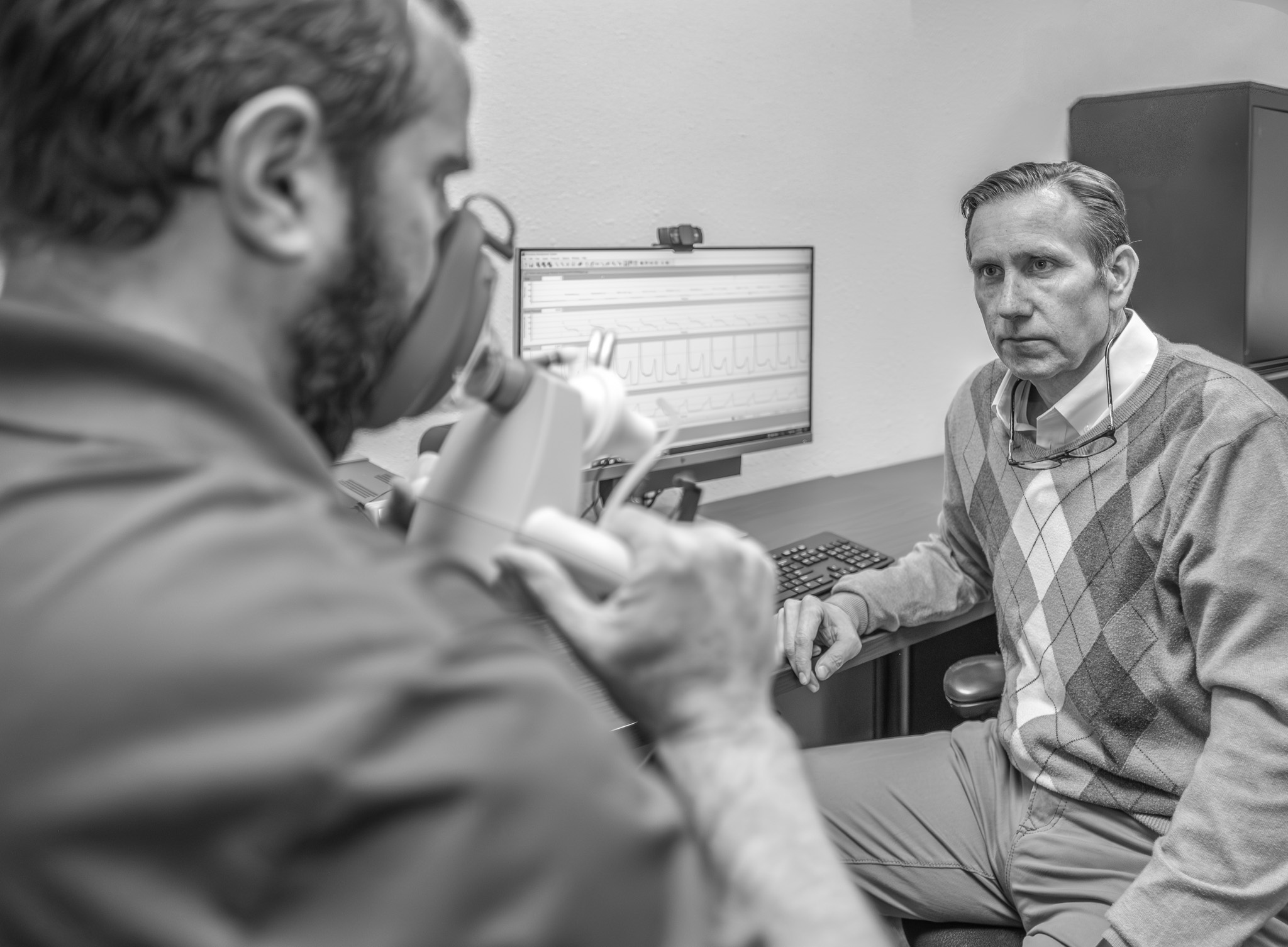 Chris Watts, wearing an argyle sweater, sits attentively in his office while Sean Taylor demonstrates handheld medical equipment. A computer monitor displaying medical data graphs is visible in the background. Black and white photo taken in the Center for Neurodegenerative Disease, TCU, December 2025.