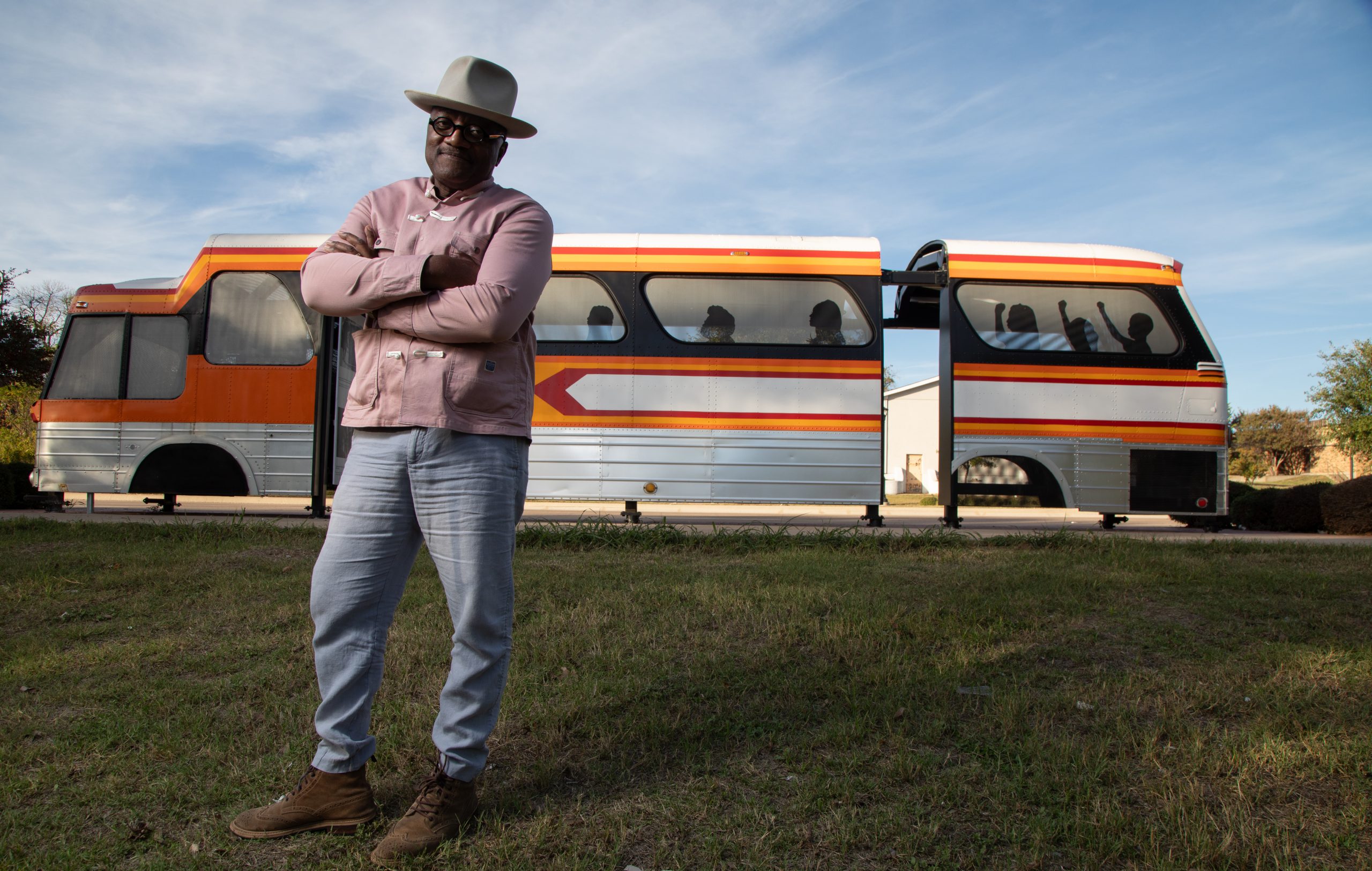 Christopher Blay, TCU alum, creator and curator, stands with arms crossed in front of a vintage orange, yellow and white striped bus, wearing a tan wide-brimmed hat, pink button-up shirt, and light jeans