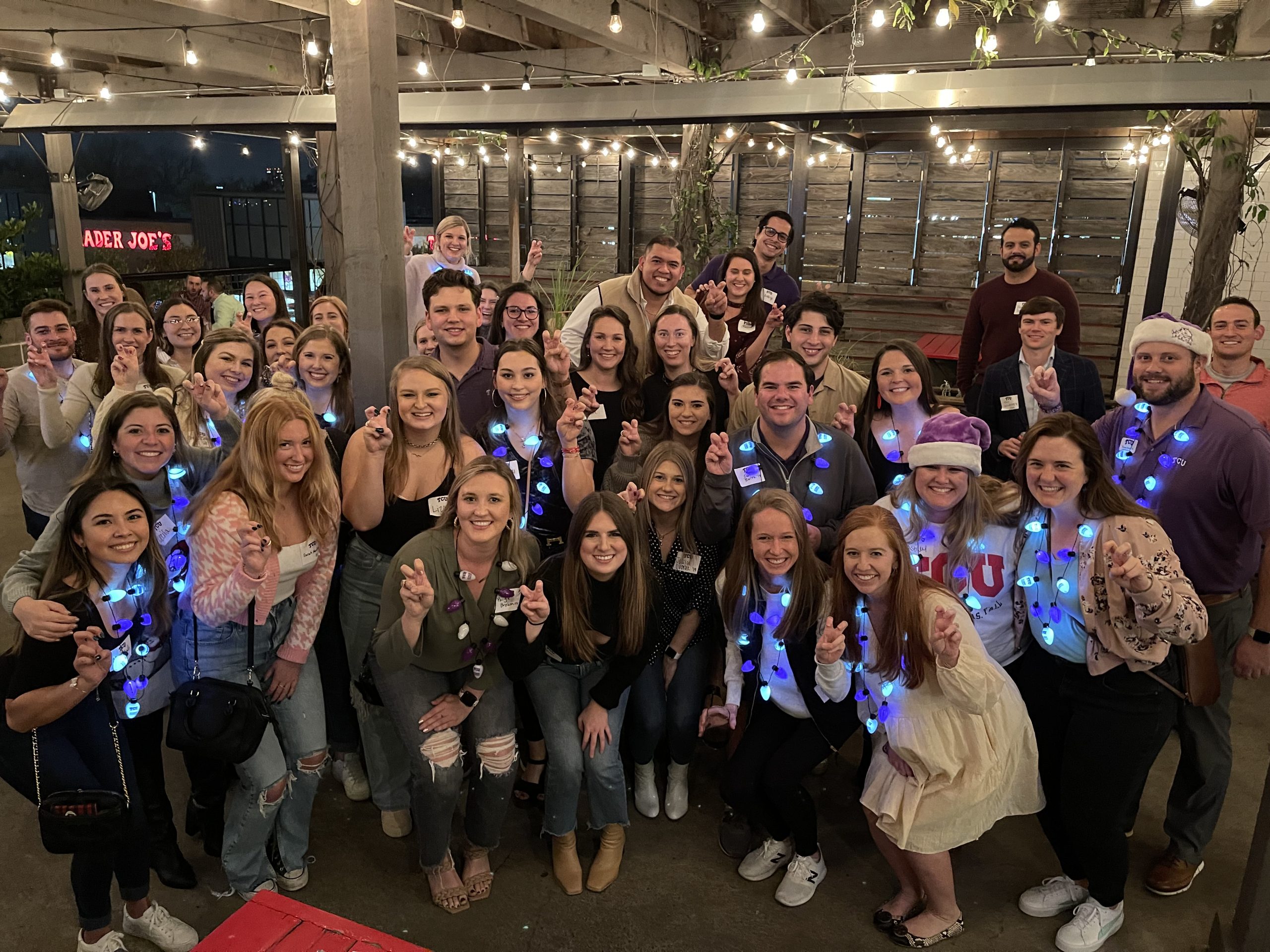 Large group of TCU young alumni holding blue LED lights and showing horns up hand signs at a holiday gathering.