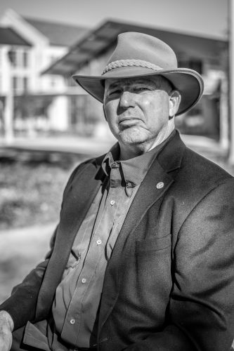 A black-and-white portrait of Dan Novak wearing a wide-brimmed hat, blazer and button-down shirt sits outdoors in front of a blurred modern building on a sunny day.