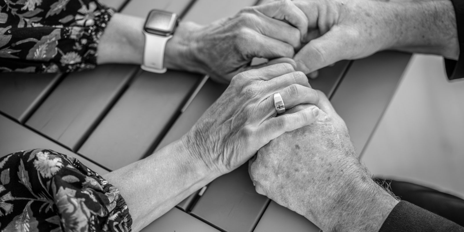 Black and white close-up of three pairs of hands clasped together over a slatted outdoor table, showing elderly hands with a cross ring and another pair of hands wearing a smartwatch and floral-patterned sleeve, conveying comfort and connection across generations.