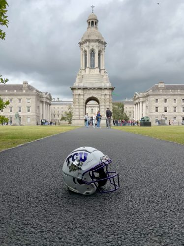 A TCU football helmet sits on a pathway in front of Trinity College Dublin's iconic Campanile bell tower, with historic stone buildings and visitors visible under dramatic cloudy skies.
