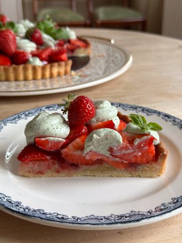 A slice of strawberry tart topped with dollops of green herb cream and a whole strawberry, served on a blue and white plate, with a full tart visible in the background.