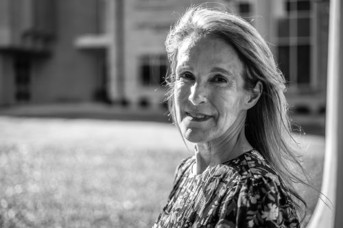Michelle Kimzey smiles outdoors on a sunny day, her long hair swept to one side, with a blurred campus building and lawn visible in the background. Black and white portrait photo.