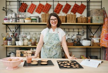 Molly Wilkinson, smiling and wearing glasses and a light green floral apron, stands behind a wooden worktable displaying two trays of baked tart shells, a bowl of eggs and a jar with a brush. Behind her, open shelving holds glass jars of ingredients, wicker baskets, baking tools and silicone molds hanging on the wall.