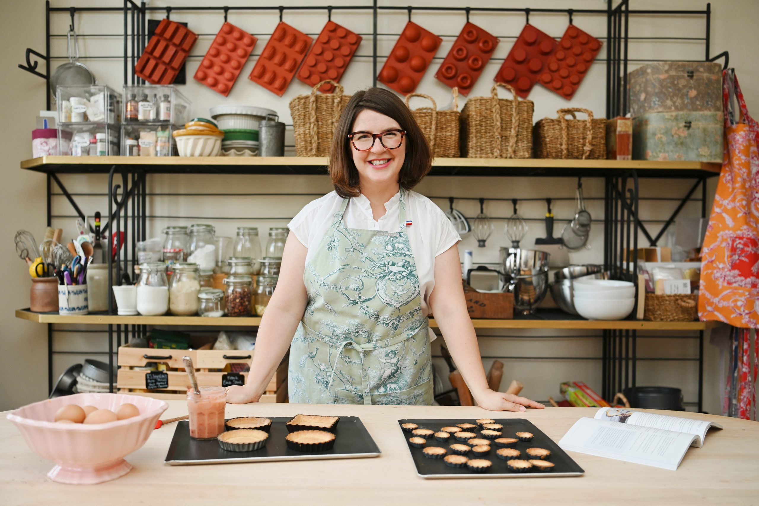 Molly Wilkinson, smiling and wearing glasses and a light green floral apron, stands behind a wooden worktable displaying two trays of baked tart shells, a bowl of eggs and a jar with a brush. Behind her, open shelving holds glass jars of ingredients, wicker baskets, baking tools and silicone molds hanging on the wall.