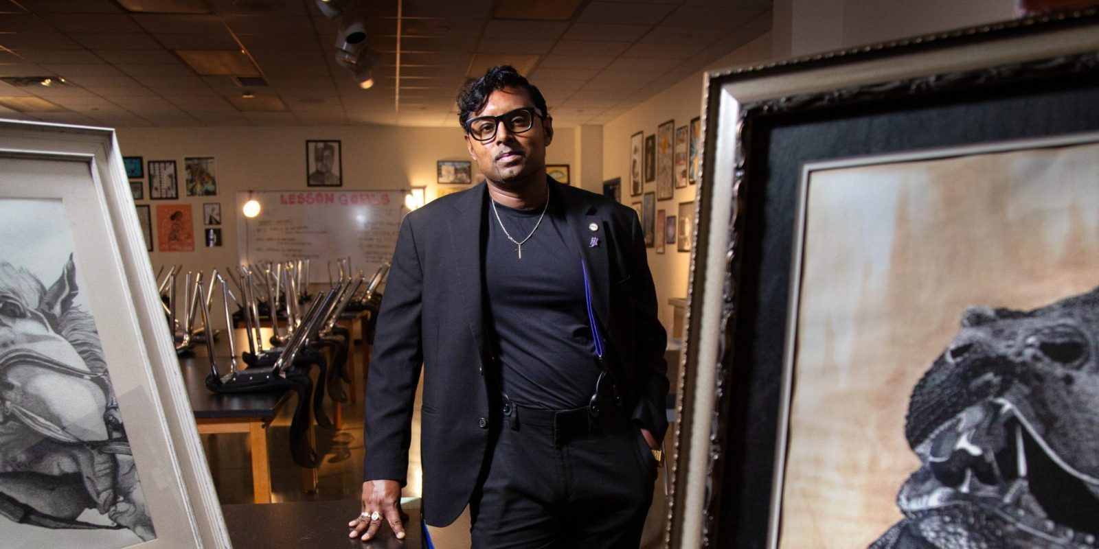DJ Perera, TCU alumna, artist, and art educator, stands between two large framed artworks in what appears to be an art classroom or gallery space. Perera, wearing a black blazer over a dark shirt with black pants and glasses, leans against a table with one hand while looking directly at the camera. Easels, artwork, and student work line the walls in the background.