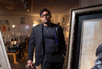DJ Perera, TCU alumna, artist, and art educator, stands between two large framed artworks in what appears to be an art classroom or gallery space. Perera, wearing a black blazer over a dark shirt with black pants and glasses, leans against a table with one hand while looking directly at the camera. Easels, artwork, and student work line the walls in the background.