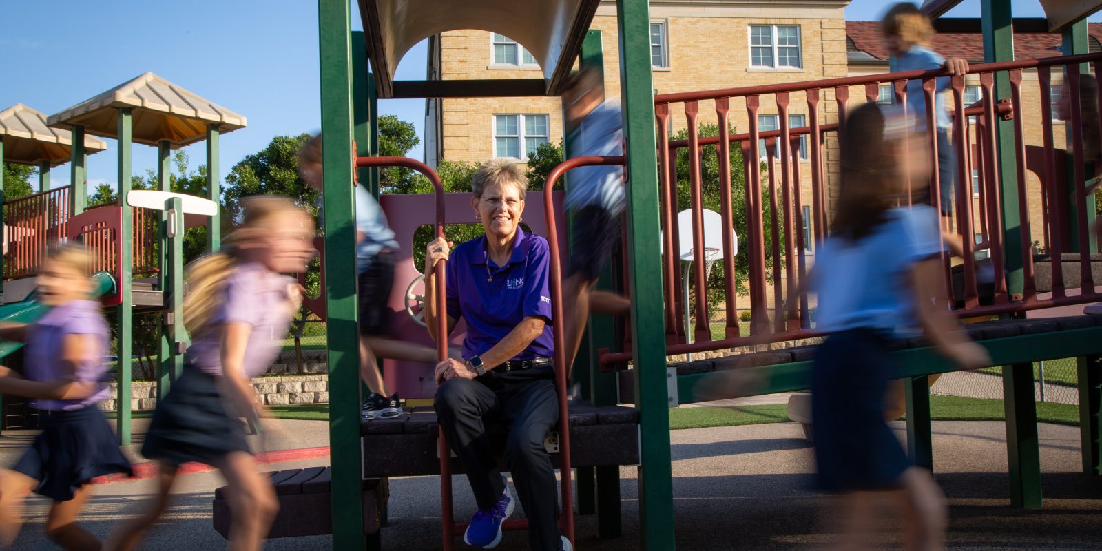 Debbie Rhea sits on playground equipment while children play around her in motion-blurred activity at a school playground.