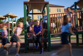 Debbie Rhea sits on playground equipment while children play around her in motion-blurred activity at a school playground.