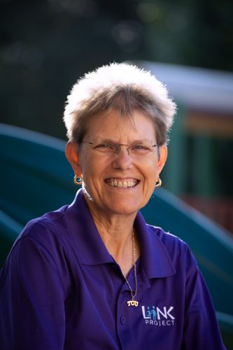 Debbie Rhea, TCU professor of kinesiology and director of the LiiNK Project, smiles at the camera wearing a purple LiiNK Project polo shirt and necklace on a school playground.