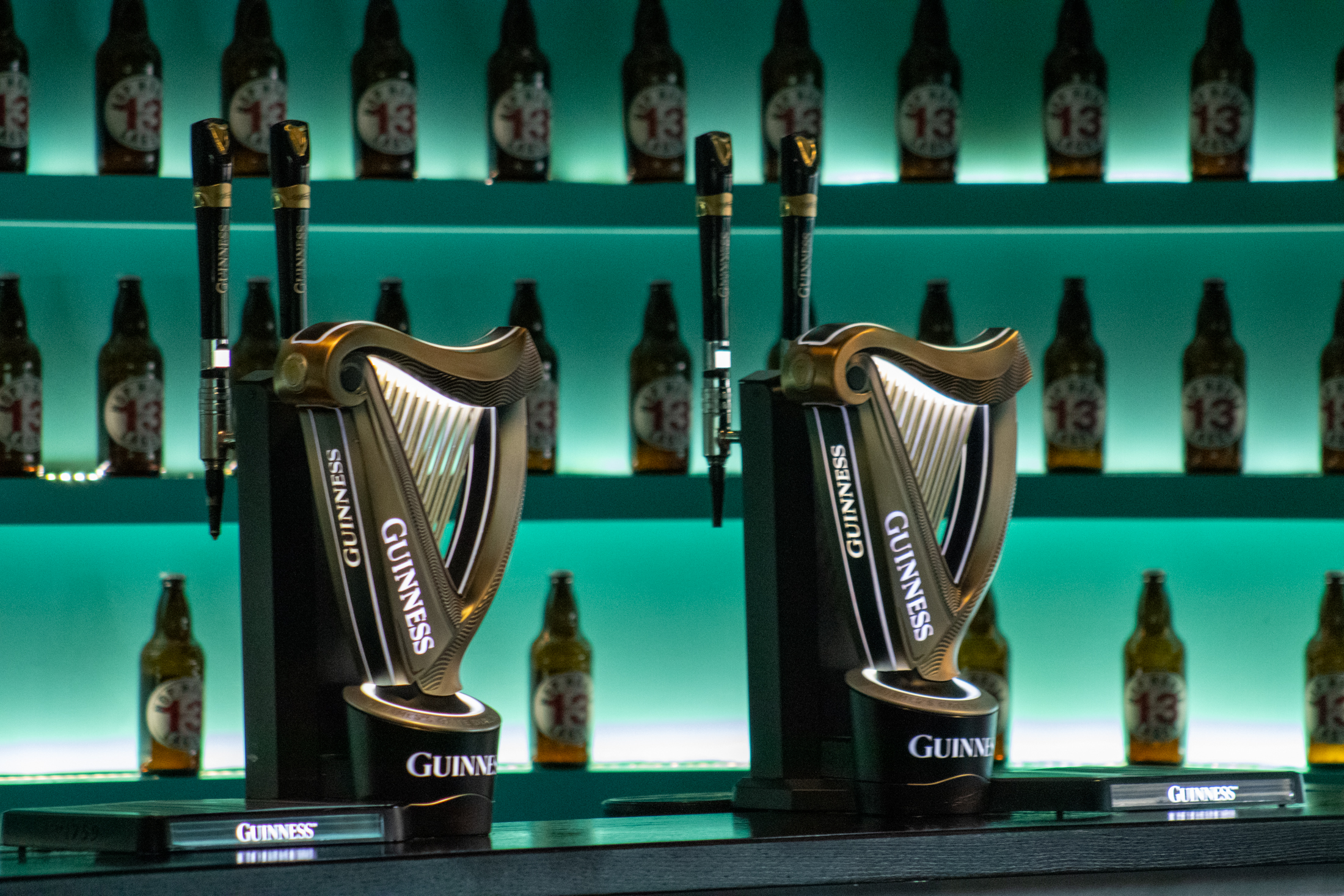 Two Guinness beer taps with the iconic harp logo stand at a bar, backed by illuminated shelves displaying rows of Guinness bottles in turquoise lighting.