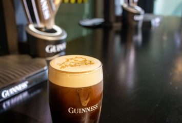 A pint of Guinness with a creamy head featuring latte art of the TCU Horned Frog mascot, sitting on a bar with additional Guinness branded glassware blurred in the background.