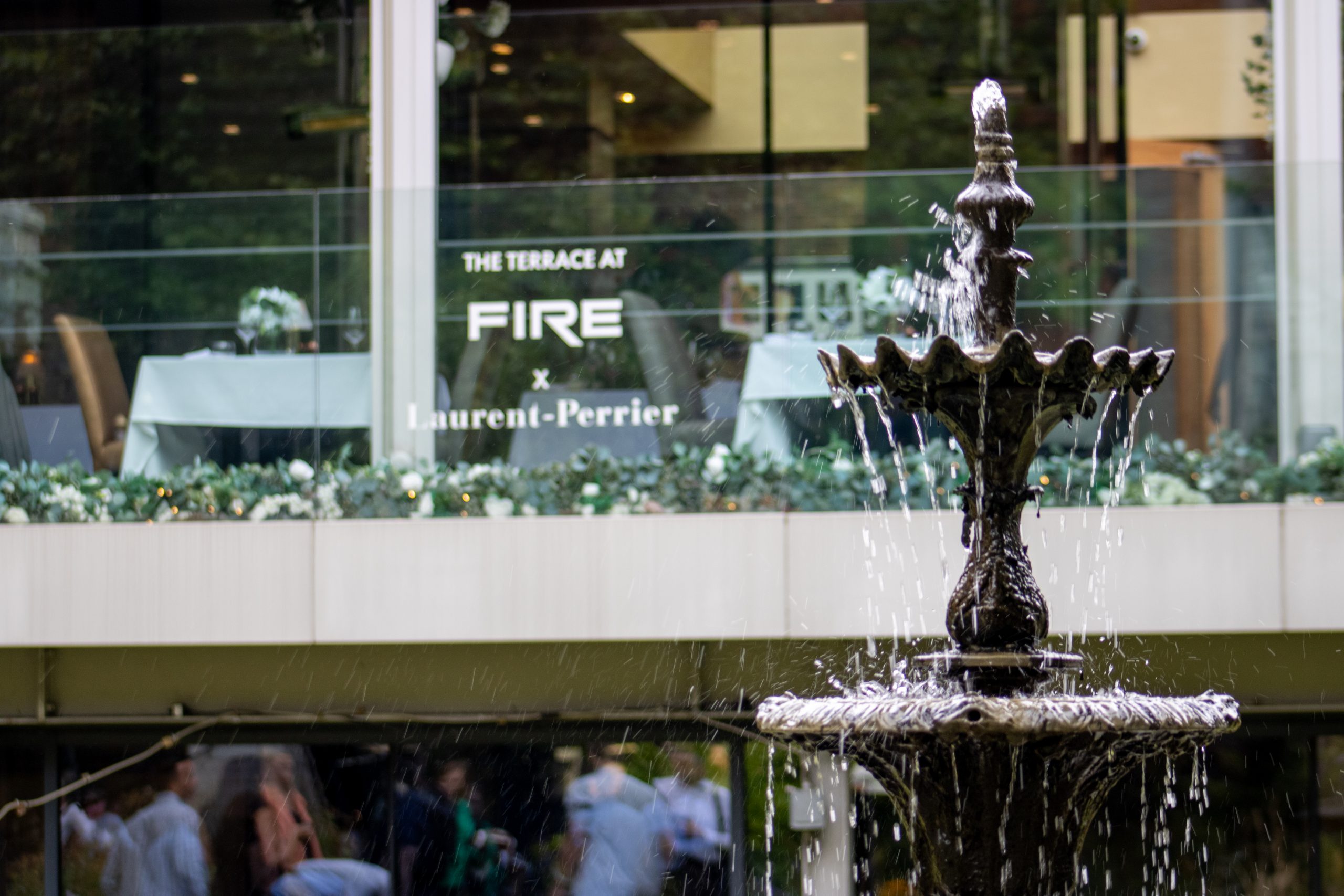 An ornate tiered fountain flows in front of "The Terrace at FIRE x Laurent-Perrier" restaurant, with glass windows revealing diners inside and landscaped planters lining the modern exterior.