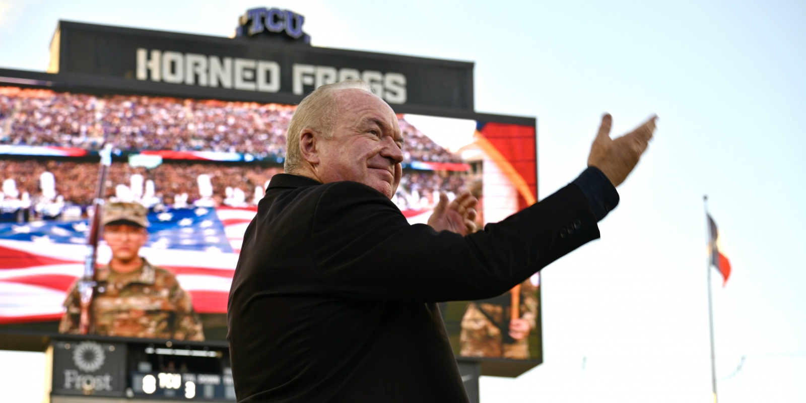 Conductor Bobby Francis gestures while leading a performance at TCU's Amon G. Carter Stadium, with an American flag display and military personnel visible on the field behind him and a packed crowd in the stands.