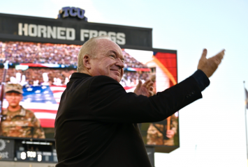 Conductor Bobby Francis gestures while leading a performance at TCU's Amon G. Carter Stadium, with an American flag display and military personnel visible on the field behind him and a packed crowd in the stands.
