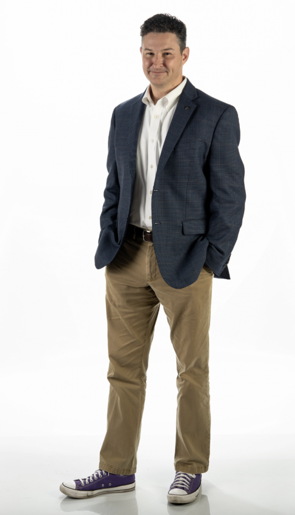 Michael Faggella-Luby, TCU College of Education professor, stands against a white background with hands in his pockets, wearing a navy blue houndstooth blazer, white button-down shirt, khaki pants, brown belt and purple Converse sneakers. He has dark curly hair and is smiling warmly at the camera in a full-length professional portrait.