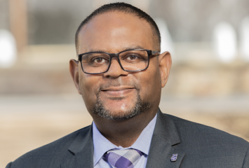Headshot of Floyd L. Wormley Jr., TCU Provost and Vice Chancellor for Academic Affairs, wearing a gray suit, light blue dress shirt, and purple plaid tie, with a pocket square and a small lapel pin, smiling warmly against a softly blurred outdoor background.