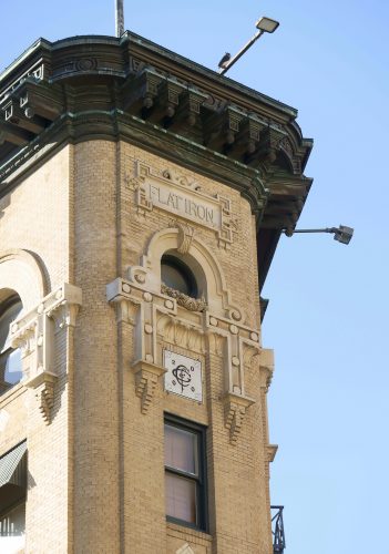 Historic Flatiron Building tower in downtown Fort Worth, showing ornate brick and terra cotta architectural details, with 'FLATIRON' carved near the top and a weathered green cornice.