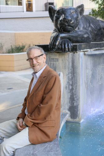 Texas historian Richard Selcer sits beside a panther sculpture atop a fountain in downtown Fort Worth, smiling at the camera while wearing a tan blazer and glasses.