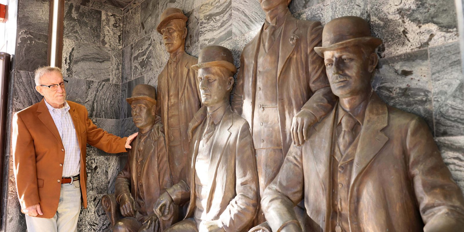 Texas historian Richard Selcer stands beside a group of bronze sculptures depicting figures in period clothing and hats, set against a rustic stone wall background.