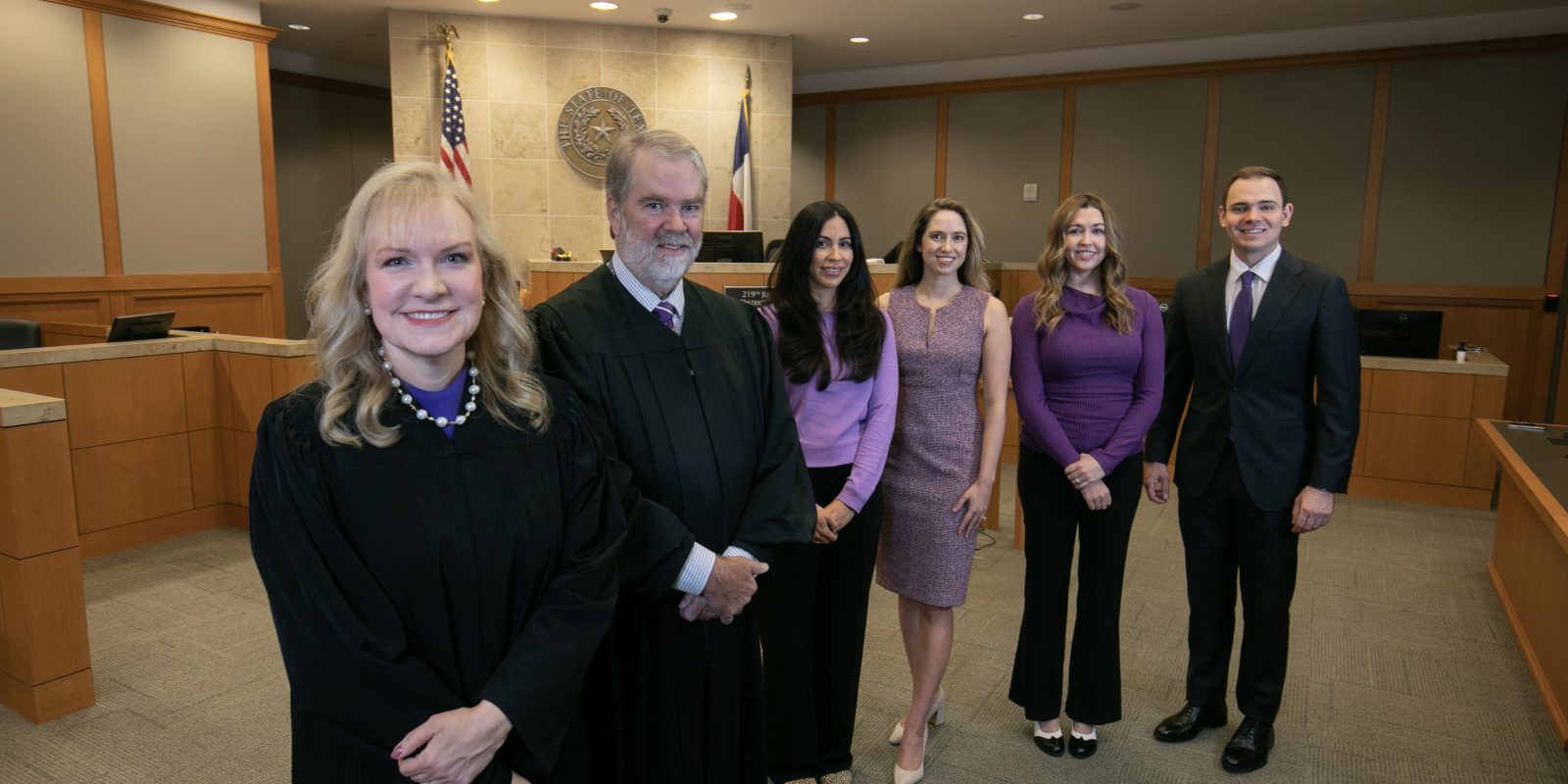 In a Collin County courtroom, judges Jennifer Edgeworth and Lance Baxter stand in the foreground wearing black robes. Behind them are four team members in business attire, from left: Michelle Garcia, Kailey Gillman, Janessa Reid and Rogan McDaniel. Edgeworth and Baxter co-founded the Collin County Adult Mental Health Court and are TCU-educated judges.