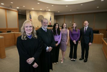 In a Collin County courtroom, judges Jennifer Edgeworth and Lance Baxter stand in the foreground wearing black robes. Behind them are four team members in business attire, from left: Michelle Garcia, Kailey Gillman, Janessa Reid and Rogan McDaniel. Edgeworth and Baxter co-founded the Collin County Adult Mental Health Court and are TCU-educated judges.