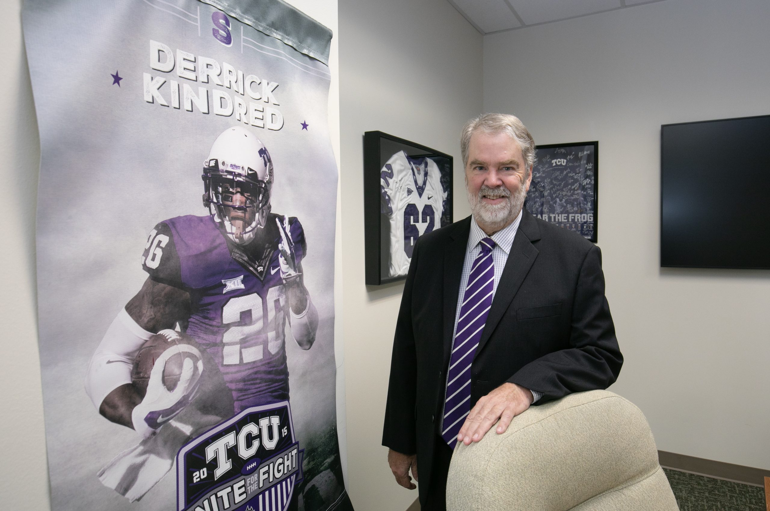 Judge Lance Baxter in his office, wearing TCU colors, with TCU football memorabilia including a Derrick Kindred poster and framed jersey displayed on the walls behind him.