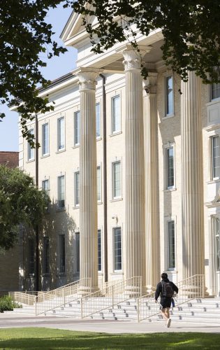 A person with a backpack walking up the steps toward a large neoclassical building with tall columns and multiple windows, framed by leafy tree branches overhead.