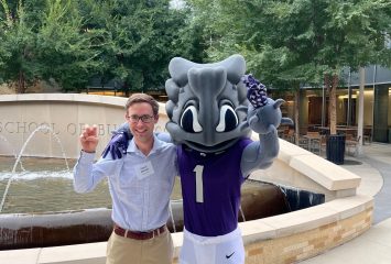 A man in business casual attire poses with SuperFrog, TCU's horned frog mascot wearing a purple jersey with number 1, in front of a fountain outside the School of Business building on campus.