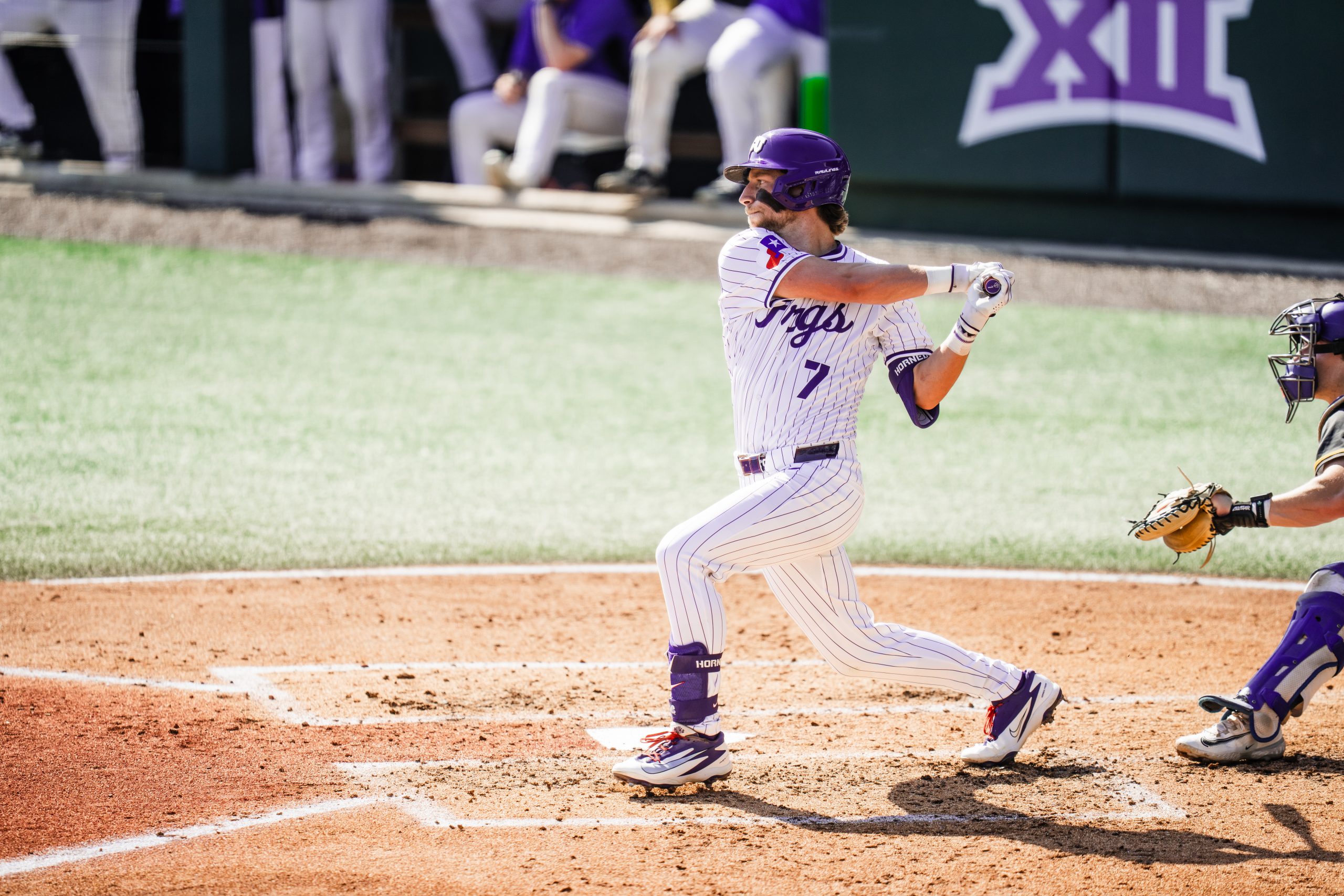 Cole Cramer follows through on a swing during a sunny afternoon home game against New Haven, wearing a white purple-pinstriped TCU Horned Frogs baseball uniform.