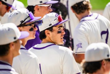 TCU baseball player Jack Bell stands among teammates, wearing a white and purple Horned Frogs uniform with eye black and red-tinted sunglasses resting on his cap.