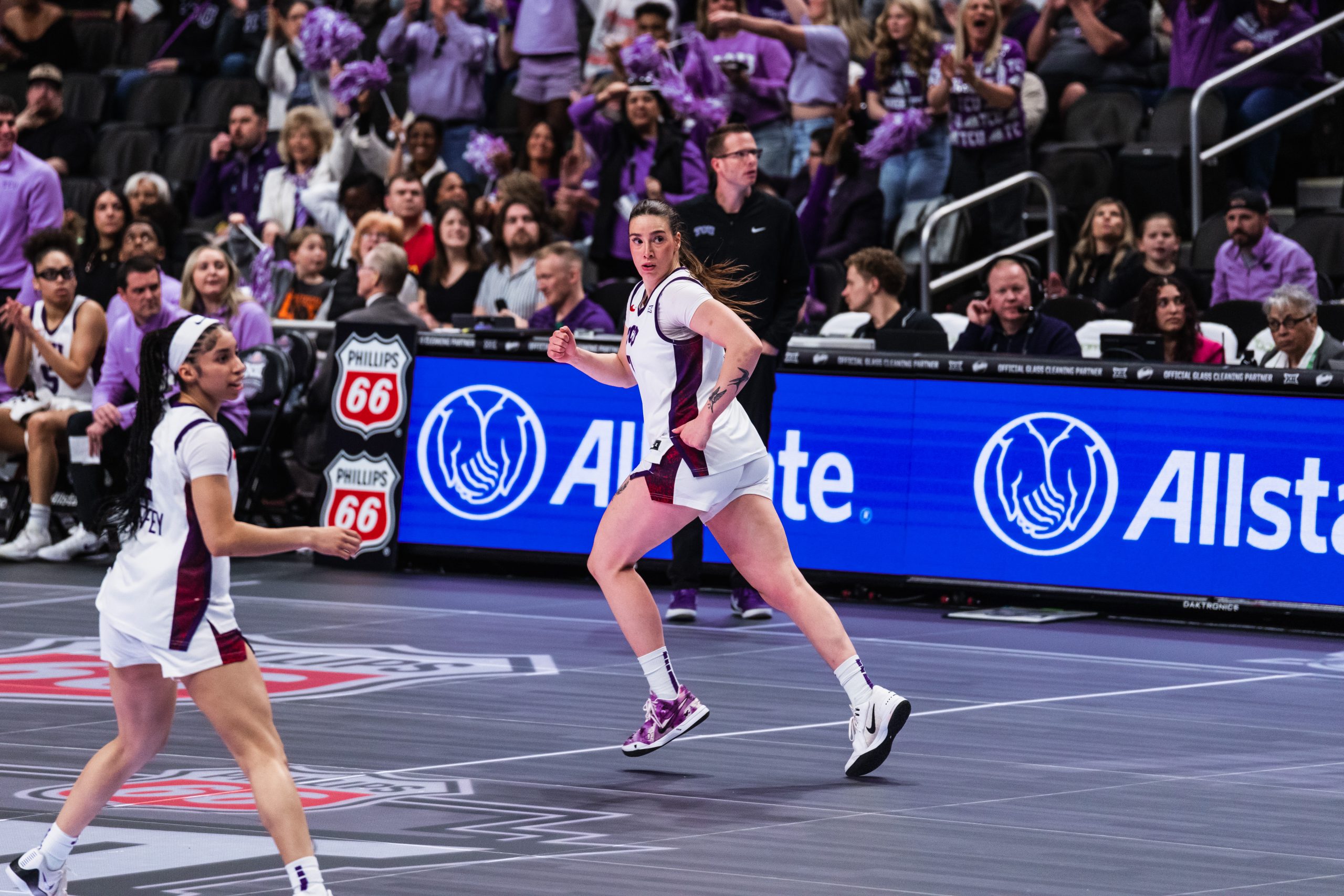 TCU forward Marta Suarez sprints up the court, her hair trailing behind her as she looks back over her shoulder. A teammate runs alongside her to the left. The crowd behind them is on its feet, cheering. Allstate and Phillips 66 branding lines the courtside boards. The game appears to be the Big 12 Tournament matchup against West Virginia.