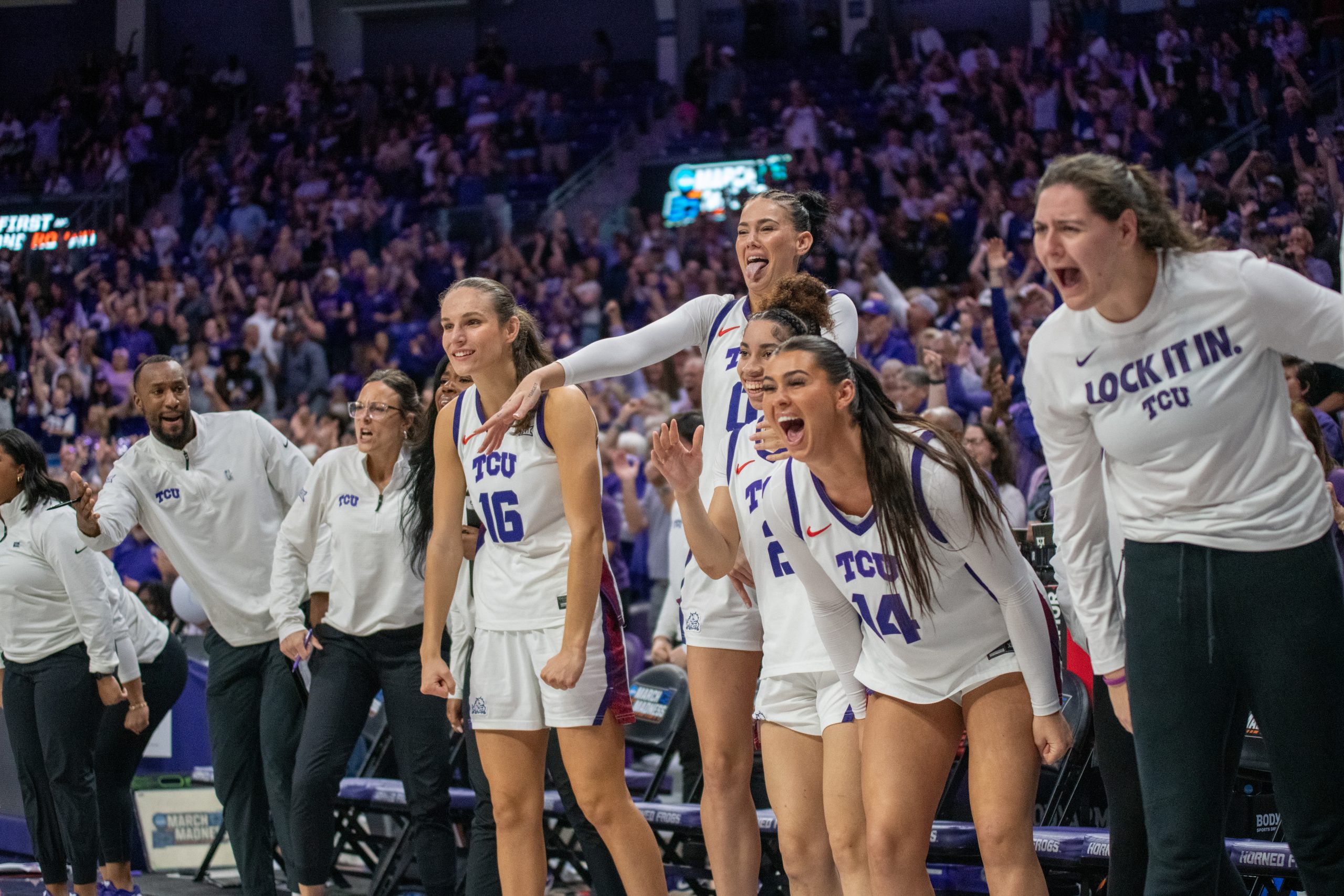 TCU women’s basketball players in white uniforms stand from the bench, cheering during a second-round NCAA Tournament game at Schollmaier Arena.