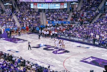 A packed Schollmaier Arena during the NCAA Women's Basketball Tournament Second Round. Two teams — TCU in purple and Louisville in red — huddle separately on the court during a stoppage in play in the fourth quarter. A referee stands at center court with one arm raised. The arena is filled with fans wearing purple, and courtside signage reads "NCAA Second Round Hosted by TCU." March Madness branding lines the court.
