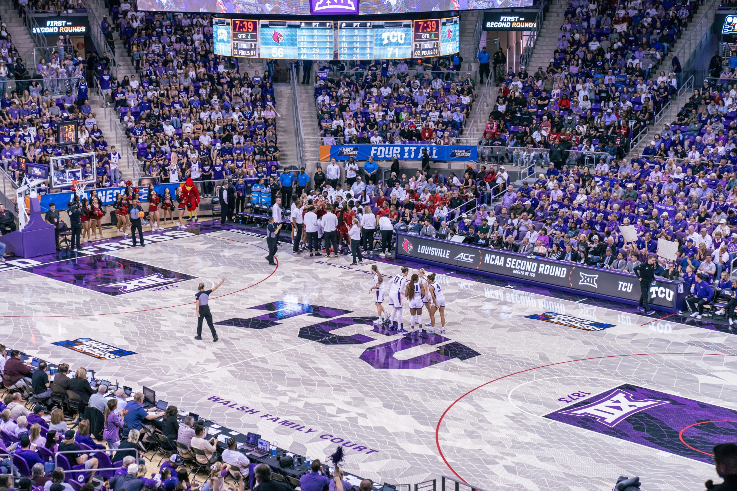 A packed Schollmaier Arena during the NCAA Women's Basketball Tournament Second Round. Two teams — TCU in purple and Louisville in red — huddle separately on the court during a stoppage in play in the fourth quarter. A referee stands at center court with one arm raised. The arena is filled with fans wearing purple, and courtside signage reads "NCAA Second Round Hosted by TCU." March Madness branding lines the court.