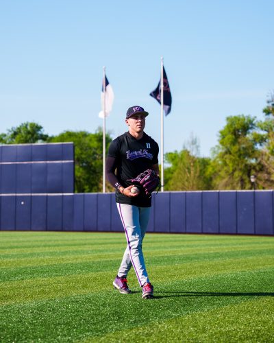 Photograph of TCU baseball player Colton Griffin in grey baseball pants with a black TCU baseball cap and top, holding a baseball in the outfield during warmups ahead of a March 2026 game at Dallas Baptist University.