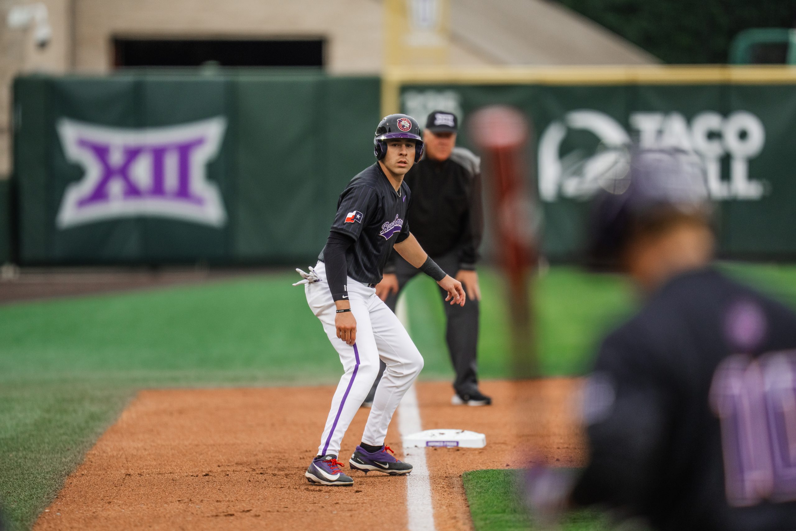 Photograph of TCU baseball player Colton Griffin during a 2026 game at Williams-Reilly Field. He stands near the third-base bag, focused on a teammate in the batter’s box, blurred in the foreground.