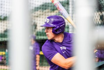 TCU baseball player Colton Griffin grips a bat during batting practice, wearing a purple helmet and shirt, seen through the blurred bars of a batting cage.
