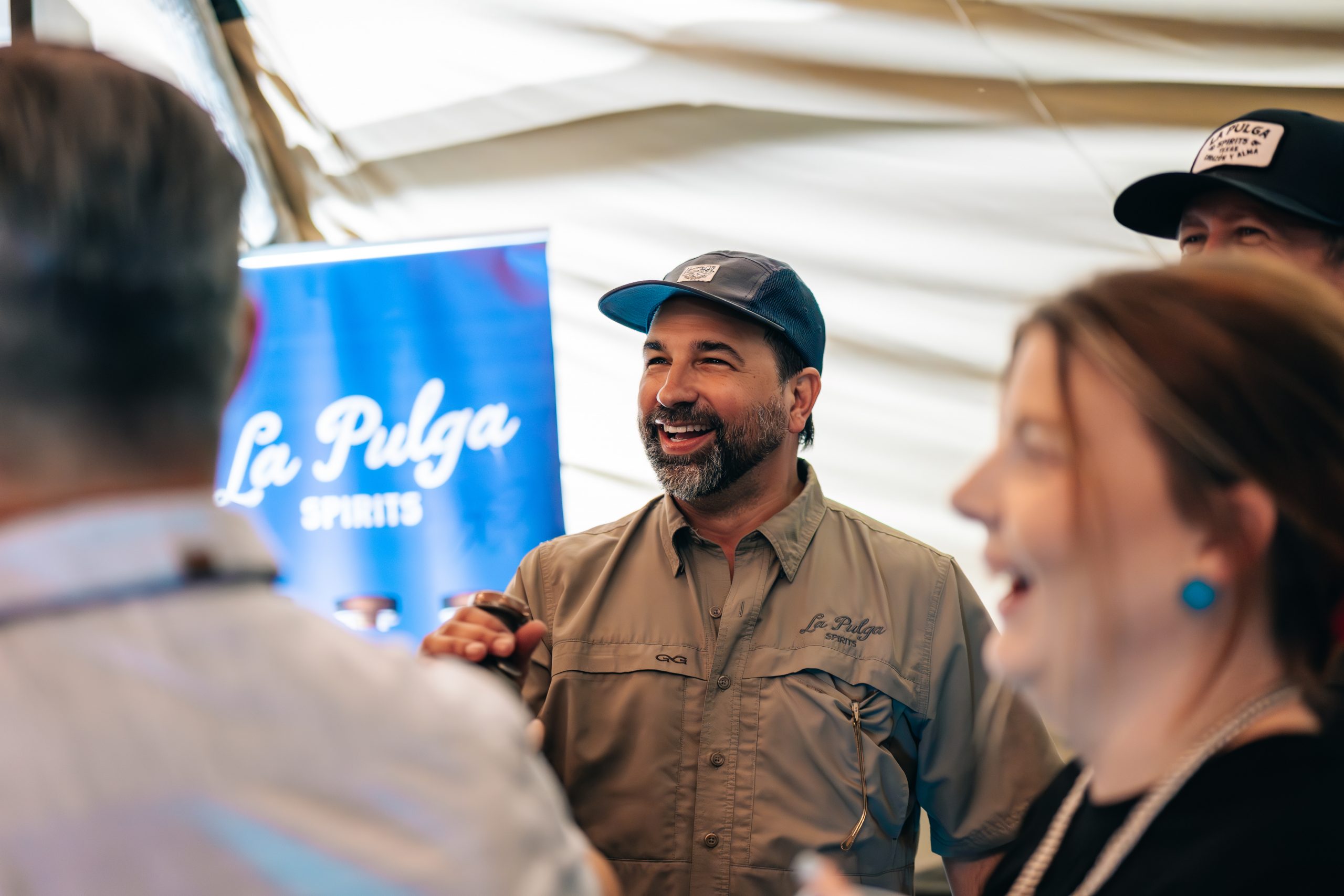 Andrew de la Torre laughs while speaking with guests at an event, with a La Pulga Spirits banner visible in the background.