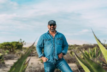 Andrew de la Torre stands among agave plants in a desert landscape, wearing a denim shirt, jeans, baseball cap and sunglasses, smiling at the camera.